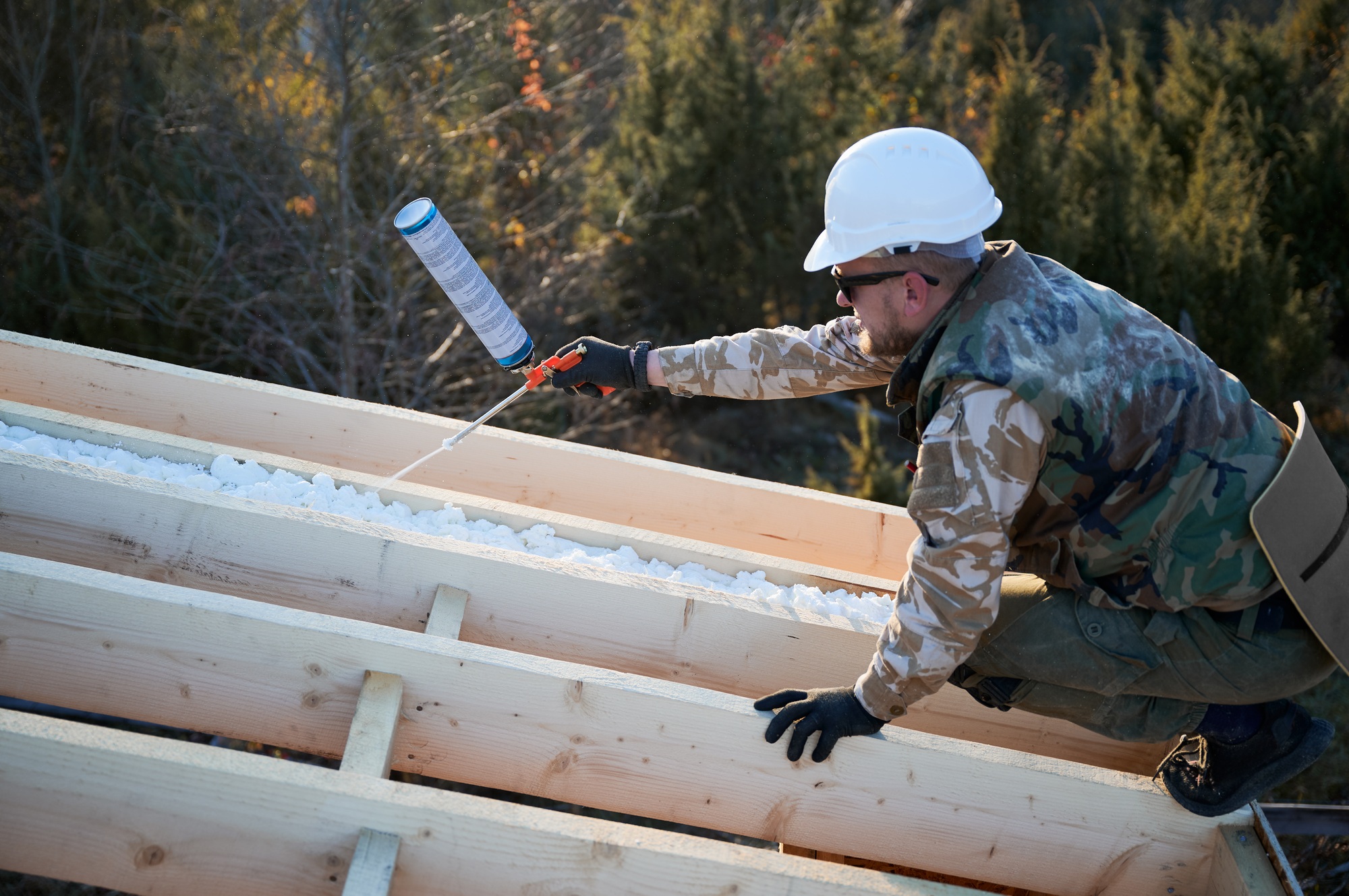 Male builder doing thermal insulation on roof of wooden frame house by polyurethane foam.